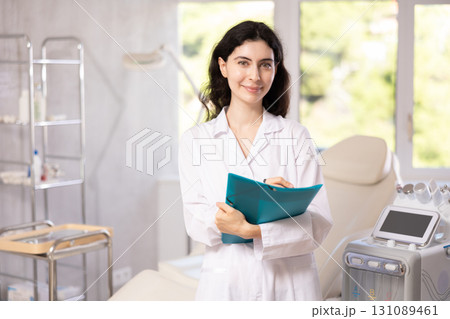 Woman doctor in uniform standing in clinic, filling out clipboard with medical records Woman doctor in uniform standing in clinic, filling out clipboard with medical records 131089461