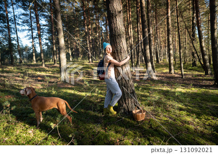 Relaxed woman hugging tree trunk in autumn forest with closed eyes at sunset, walk with dog in woods 131092021