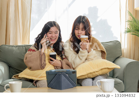 Winter day brings women friends together at home, enjoying cozy friendship indoors, talking on phone, watching tablet with warm coffee, embracing comforting winter leisure activities. Winter day brings women friends together at home, enjoying cozy friendship indoors, talking on phone, watching tablet with warm coffee, embracing comforting winter leisure activities. 131094832