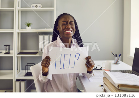 Stressed woman sitting at her workplace in office holding a sign with the inscription word help. Stressed woman sitting at her workplace in office holding a sign with the inscription word help. 131095116
