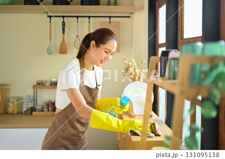 Smiling woman in apron and gloves cleaning wooden kitchen counter with spray bottle and cloth 131095138