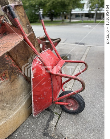 A Red Wheelbarrow is Leaning Against a Wall in a Busy Urban Setting or Environment 131095464