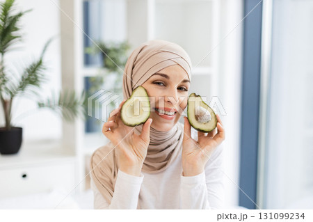 Smiling young Muslim woman holding avocado halves, wearing hijab in modern bright interior setting 131099234