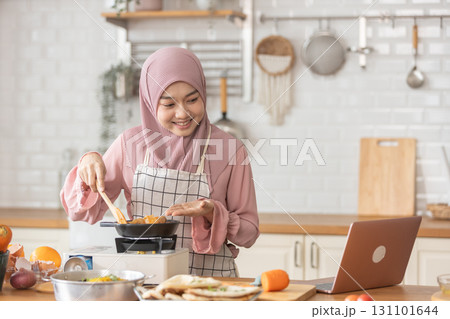 Muslim woman cooking food in modern kitchen while watching recipe on laptop. Wearing hijab and apron, preparing meal with fresh ingredients. Concept of halal food, home cooking, and lifestyle. 131101644