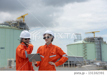Two engineers in orange uniforms and safety helmets discussing project data at a gas plant with digital tablet and radio, representing teamwork, technology, energy industry, and field operations. 131102391