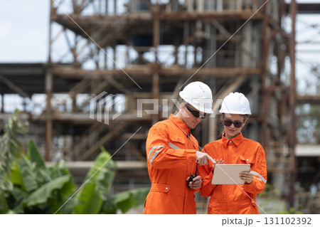 Two engineers in orange protective uniforms and white helmets discussing industrial site plans using a digital tablet, focusing on construction safety, teamwork, and technical coordination. 131102392
