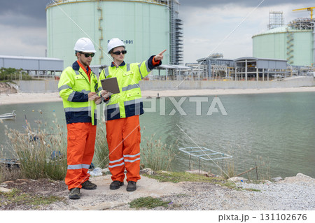 Two engineers wearing high-visibility safety jackets and helmets standing by the water, discussing inspection plans and pointing toward a large industrial gas storage facility in the background. Two engineers wearing high-visibility safety jackets and helmets standing by the water, discussing inspection plans and pointing toward a large industrial gas storage facility in the background. 131102676