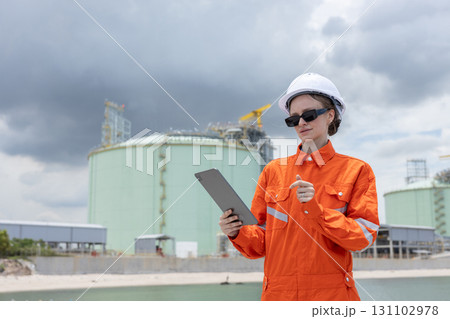 Female engineer in orange safety uniform and white helmet using a tablet while inspecting an industrial gas storage facility, representing technology, safety, and energy industry operations. 131102978