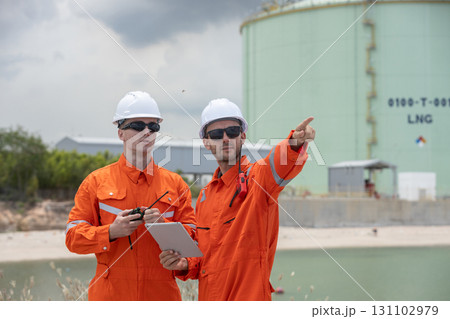 Two male engineer in orange protective uniforms and white helmets discussing site operations at LNG gas plant with a tablet and walkie-talkie, emphasizing energy industry, safety, and teamwork. Two male engineer in orange protective uniforms and white helmets discussing site operations at LNG gas plant with a tablet and walkie-talkie, emphasizing energy industry, safety, and teamwork. 131102979