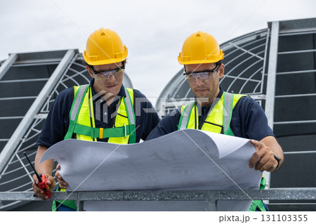 Two engineers in safety helmets and vests examine a construction blueprint on a rooftop HVAC site. Concept of industrial planning, project management, and mechanical system inspection. Two engineers in safety helmets and vests examine a construction blueprint on a rooftop HVAC site. Concept of industrial planning, project management, and mechanical system inspection. 131103355