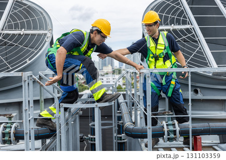 Two engineers wearing safety gear assist each other while crossing rooftop HVAC pipes. Concept of teamwork, industrial safety, facility maintenance, and support in hazardous work environments. Two engineers wearing safety gear assist each other while crossing rooftop HVAC pipes. Concept of teamwork, industrial safety, facility maintenance, and support in hazardous work environments. 131103359