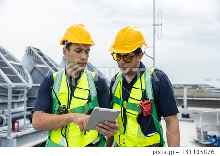 Engineers wearing safety vests and helmets discuss HVAC system inspection on rooftop. Industrial maintenance and building ventilation system operation concept. Engineers wearing safety vests and helmets discuss HVAC system inspection on rooftop. Industrial maintenance and building ventilation system operation concept. 131103876