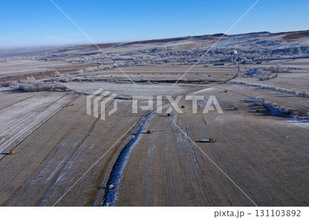 Aerial view of snow-covered field dotted with hay bales under a clear blue sky during winter Aerial view of snow-covered field dotted with hay bales under a clear blue sky during winter 131103892
