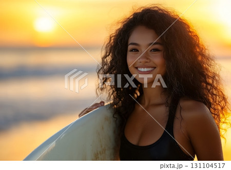 A smiling young woman with curly hair enjoys a beautiful sunset at the beach, holding her surfboard 131104517