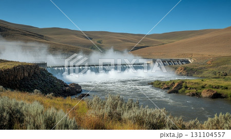 A powerful dam releases water, creating a misty spectacle over a rushing river in a scenic valley 131104557