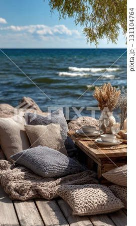 Cozy picnic by the sea. Table with tableware, soft pillows, and knit blanket create a relaxing beach getaway 131104764