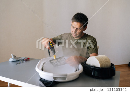 Portrait of young man using steam cleaner to clean air humidifier filter in basin at home, ensuring regular maintenance for maintaining healthy indoor air quality. Concept of domestic work. 131105542