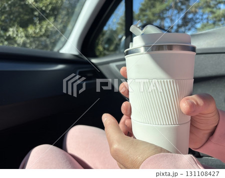 A woman holds a thermal cup with a hot drink, coffee or tea, in the car. Drinking from a travel mug during a long road trip, warming up on the way, autumn and winter travel lifestyle A woman holds a thermal cup with a hot drink, coffee or tea, in the car. Drinking from a travel mug during a long road trip, warming up on the way, autumn and winter travel lifestyle 131108427