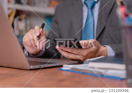 Man holding smartphone and pen, working on desk with laptop and documents, perfect for business tech, remote work, and productivity themes. Man holding smartphone and pen, working on desk with laptop and documents, perfect for business tech, remote work, and productivity themes. 131109306