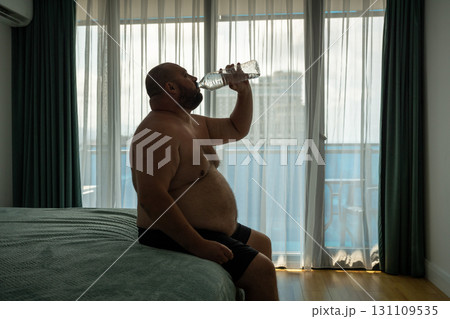 Oversize guy drinks lot of water sits on bed, trying to cool off resist heat cope with dehydration 131109535