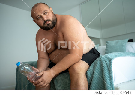 Tired fat man sits on bed with water bottle after training for weight loss think about goal future Tired fat man sits on bed with water bottle after training for weight loss think about goal future 131109543