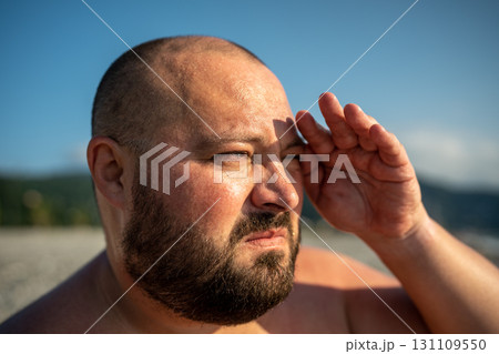 Bearded overweight man looking at distance on nature at sunny weather covering eyes from sun. 131109550