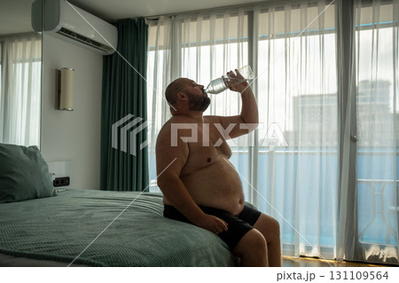 Overweight man drinking water sitting in bed in hotel room on vacation in summer hot weather. Overweight man drinking water sitting in bed in hotel room on vacation in summer hot weather. 131109564