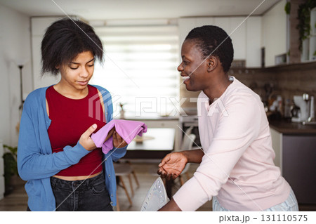Mother and daughter ironing clothes and smiling at home 131110575