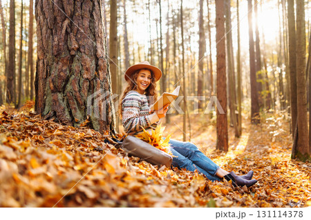 Pretty young woman in hat reading interesting book near tree in autumn forest. Enjoying nature Relax 131114378
