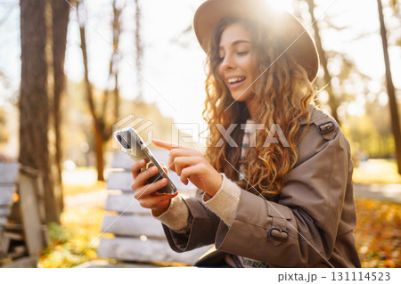 Young woman in hat on bench with telophone enjoys autumn weather in sunny park. Concept lifestyle. Young woman in hat on bench with telophone enjoys autumn weather in sunny park. Concept lifestyle. 131114523