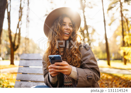 Young woman in hat on bench with telophone enjoys autumn weather in sunny park. Concept lifestyle. Young woman in hat on bench with telophone enjoys autumn weather in sunny park. Concept lifestyle. 131114524