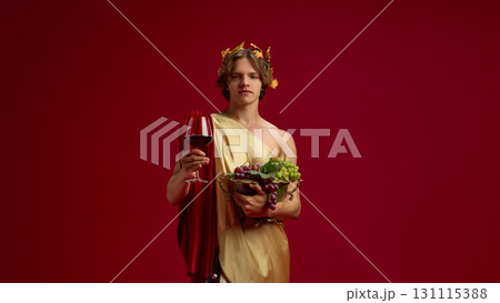 Young man in toga holding grapes and red wine glass on festive background 131115388