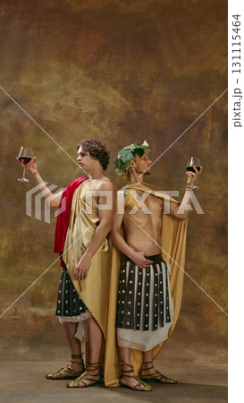 Mature man and his young student in medieval costumes standing back to back with wine glasses raised high 131115464