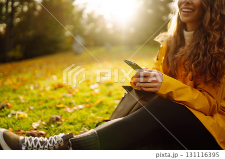 A smiling woman sits outdoors on the lawn among yellow autumn leaves with a phone in her hands. 131116395