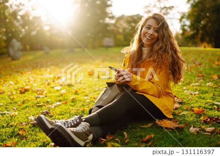 A smiling woman sits outdoors on the lawn among yellow autumn leaves with a phone in her hands. 131116397