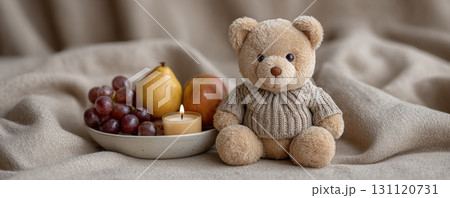 Cozy teddy bear in sweater sits beside plate of fruits and lit candle, creating warm and inviting scene 131120731