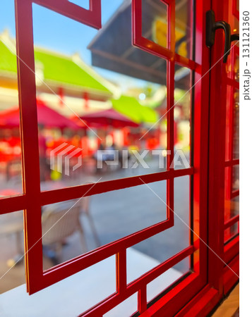 View through red lattice window into a sunny courtyard with green-tiled roofs and red umbrellas. Traditional Chinese architecture in vibrant outdoor setting. View through red lattice window into a sunny courtyard with green-tiled roofs and red umbrellas. Traditional Chinese architecture in vibrant outdoor setting. 131121360