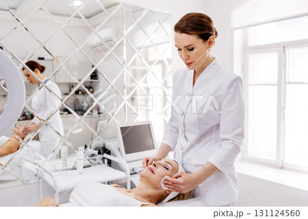 Beautician performing facial skincare treatment in modern beauty clinic. Professional cosmetologist cleaning mature woman face with cotton pads 131124560