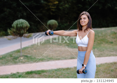 Young woman exercising with dumbbells in a park, promoting healthy lifestyle and fitness 131124945