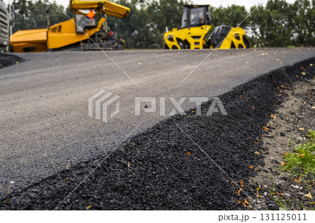 Fresh asphalt layer close-up with road construction machinery in background Fresh asphalt layer close-up with road construction machinery in background 131125011