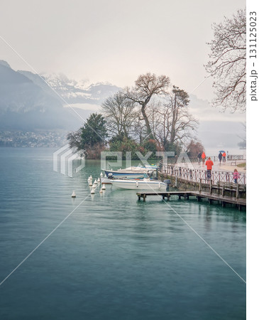 Misty lakeside scene in Annecy, France with view to the swan island. People walking the alley by the calm, turquoise water during a tranquil rain. Snow capped mountains loom in the foggy background 131125023