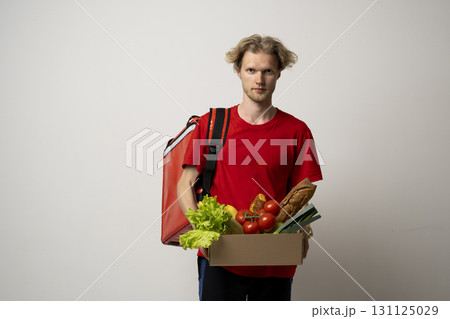 Delivery man with red insulated backpack holding grocery box full of food 131125029