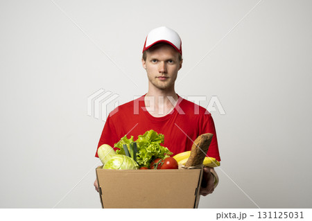 Courier in red uniform holding grocery box with fresh vegetables and bread 131125031