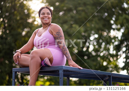 African American woman in pink sportswear sitting outdoors 131125166