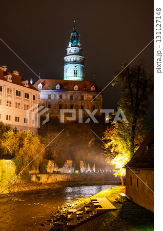 A stunning night view of the illuminated tower of Cesky Krumlov Castle towering above the Vltava River, creating a magical atmosphere with the reflection in the water 131127148