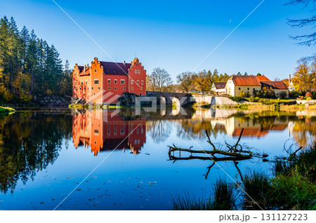 Panoramic view of Cervena Lhota Castle in the Czech Republic, surrounded by a lake reflecting its image. Autumn trees in vibrant colors and sunny weather create a picturesque, serene scene. 131127223