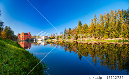 Panoramic view of Cervena Lhota Castle in the Czech Republic, surrounded by a lake reflecting its image. Autumn trees in vibrant colors and sunny weather create a picturesque, serene scene. 131127224