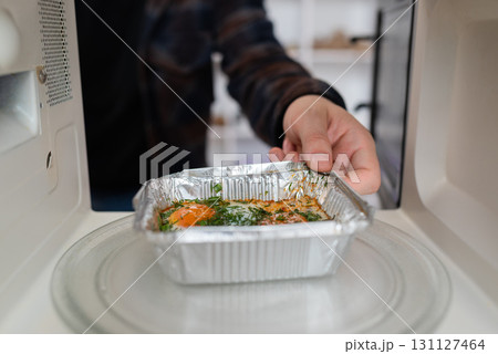 A person's hand places an aluminum takeaway container with food into a microwave, a common kitchen mistake and safety issue. A person's hand places an aluminum takeaway container with food into a microwave, a common kitchen mistake and safety issue. 131127464