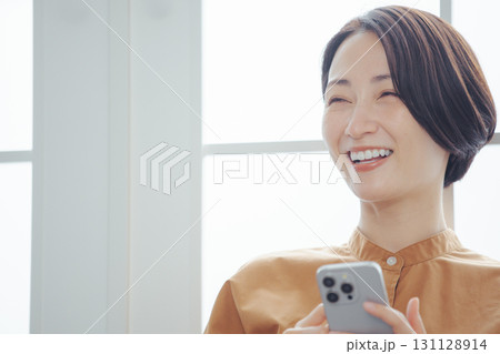 窓際でスマホを見ながら嬉しそうに微笑むミドル女性 窓際でスマホを見ながら嬉しそうに微笑むミドル女性 131128914