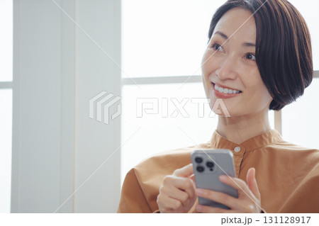 窓際でスマホを見ながら嬉しそうに微笑むミドル女性 131128917